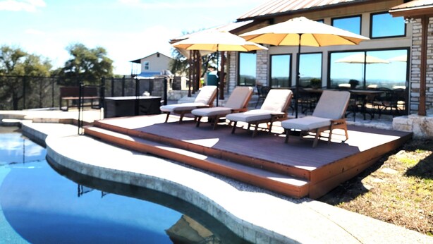 A poolside area with lounge chairs and umbrellas, a stone building with large windows, and a clear sky. - VIEWS! Texas Hill Country - Bella Vista (Dripping Springs)
