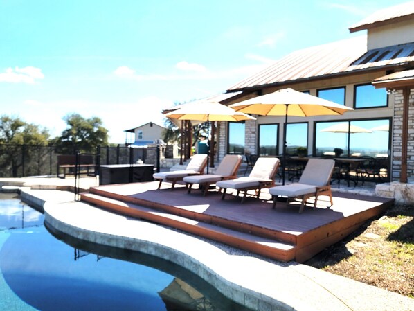 Zona de piscina con sillones de descanso y sombrillas, un edificio de piedra con grandes ventanales y cielo despejado.