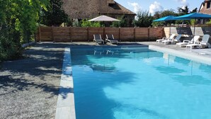 A rectangular swimming pool with clear blue water, surrounded by a wooden fence and a stone wall. There are lounge chairs and umbrellas along the poolside.