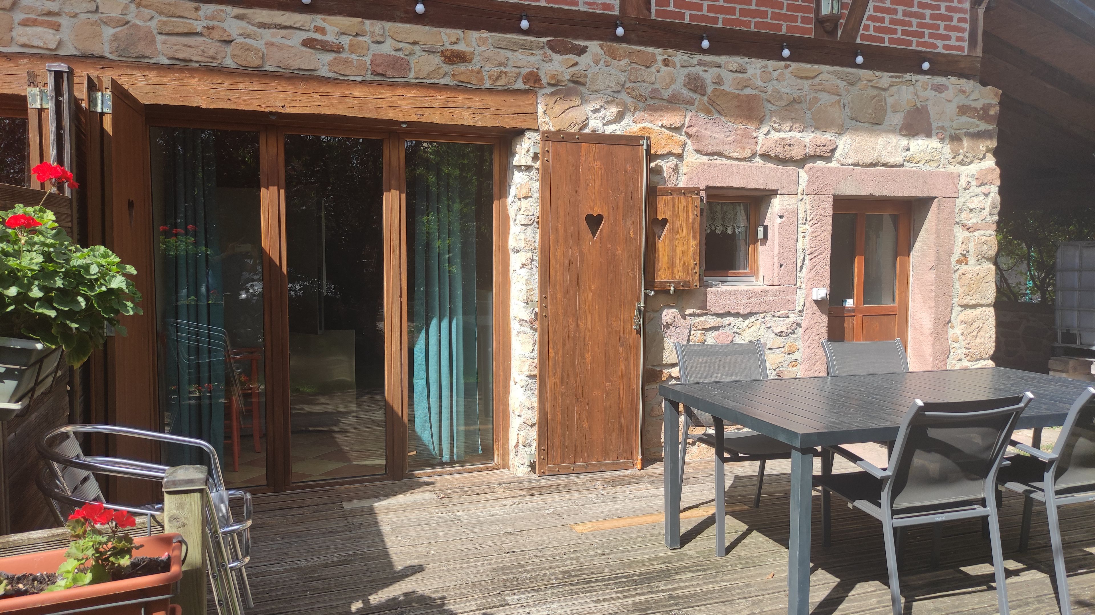 A stone building with a wooden door, a patio with a table and chairs, and a potted plant with red flowers.