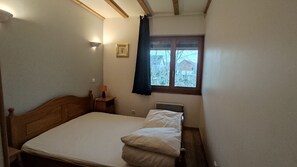 Ett litet rum med en enkel säng, ett sängbord med en lampa, ett fönster med utsikt över träd och ett hus samt en inramad tavla på väggen.