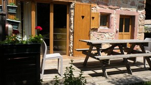 A rustic stone building with a wooden door and windows, a wooden deck with a picnic table and chairs, and potted plants with flowers.