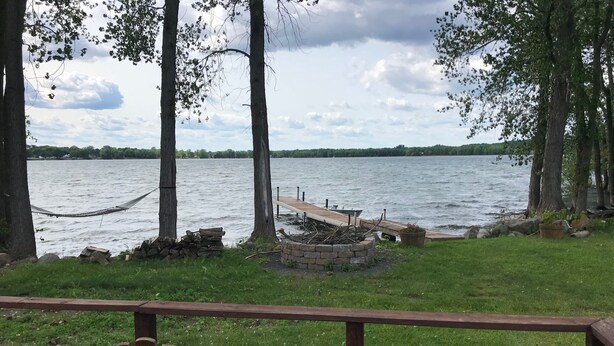 A deck with wooden chairs, a hammock, a dock, and a stone fire pit by a lake.