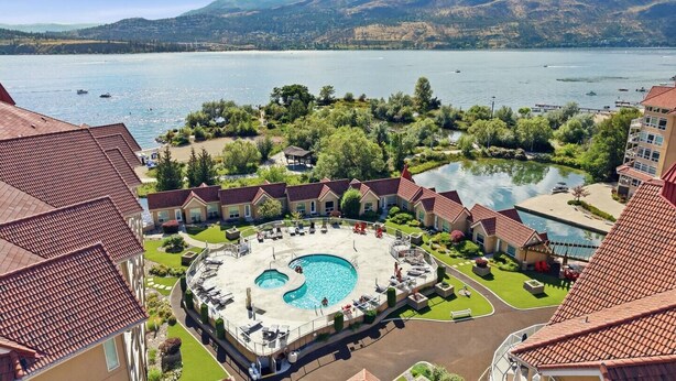 Un hotel con piscina centrale, diversi edifici dai tetti in tegole rosse e vista su un lago con barche.