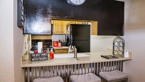 Cocina compacta con un reloj negro, un refrigerador, un lavamanos y dos taburetes.