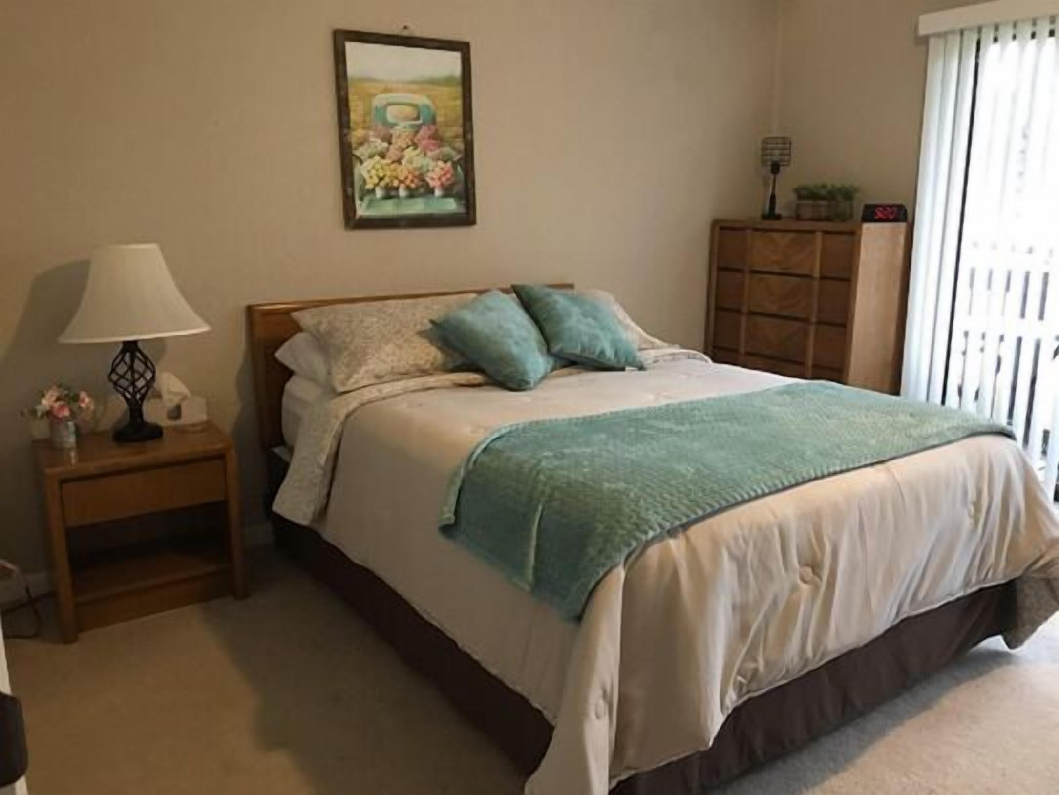 A bedroom with a bed, a nightstand, a lamp, a painting, and a window with curtains.