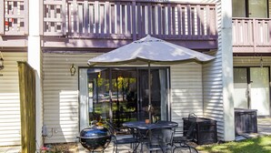 Un patio con una parrilla, mesa exterior, sillas y un paraguas grande.