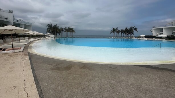 A large swimming pool with lounge chairs and umbrellas, surrounded by palm trees and a view of the ocean. - Ocean and swimming front pool view, 6 persons, 2 bedrooms and 2 bathrooms (Bahia De Banderas, Nuevo Vallarta, Bucerias)