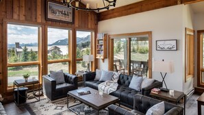 Un salon chaleureux avec un canapé en cuir, une table basse et une bibliothèque. De grandes fenêtres offrent une vue sur les montagnes et donnent sur une terrasse en bois à l’extérieur.