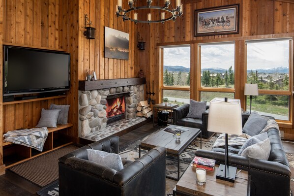 A cozy living room with a stone fireplace, leather furniture, a flat-screen TV, and large windows offering a mountain view. - Outpost: Nez Perce D6 - Hot Tub Access! (Teton Village)