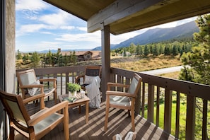 Una terraza de madera con mobiliario exterior, una mesa con una planta y una taza, y una vista panorámica de montañas.