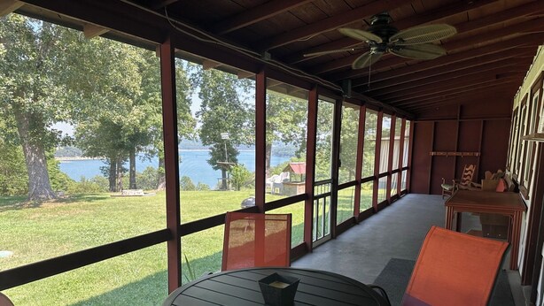 En overdækket terrasse med et bord og stole, store vinduer med udsigt over en sø og grøn vegetation.