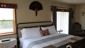 A neatly made bed with a quilt, a small bedside table with a book, and a chair in a room with a window and curtains.