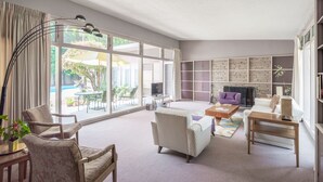 A spacious living room with a large window, a fireplace, a bookshelf, and a dining area with outdoor furniture. - Bixby Beauty 50s Mod -- Pool and Parking (Long Beach)