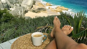 A view of a rocky coastline with clear blue water, a white cup of coffee on a woven mat, and a person's feet resting on a wooden deck. - Spectacular Eco Cottage, oceanfront, secret beach (Gregorytown)