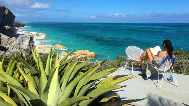 A person reading a book on a chair overlooking a coastal area with clear blue water and rocky cliffs. - Spectacular Eco Cottage, oceanfront, secret beach (Gregorytown)