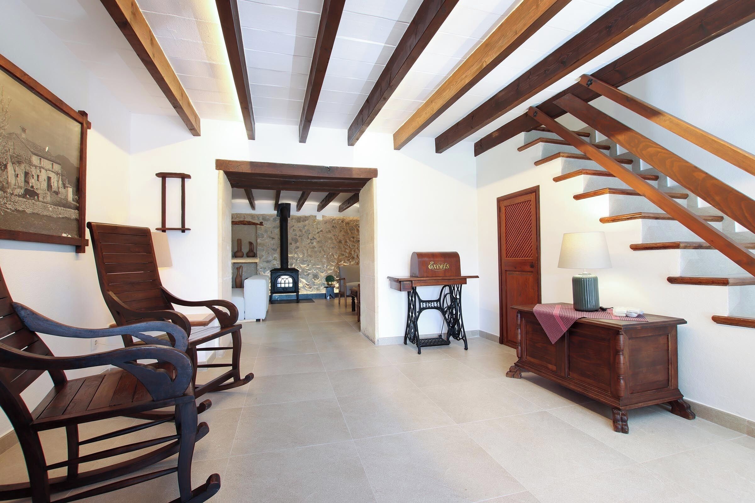 A spacious hallway with wooden beams, a staircase, rocking chairs, a sewing machine, and a lamp.