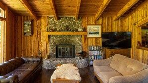 L’intérieur chaleureux d’un chalet avec une cheminée en pierre, des canapés en cuir, un téléviseur et une bibliothèque.