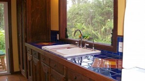 A kitchen with a double sink, wooden cabinets, and a window with a view of greenery. -  Enchanting Rainforest Hideaway, on 2 Private Acres to yourself.  Pahoa/Keaau   (Pahoa)