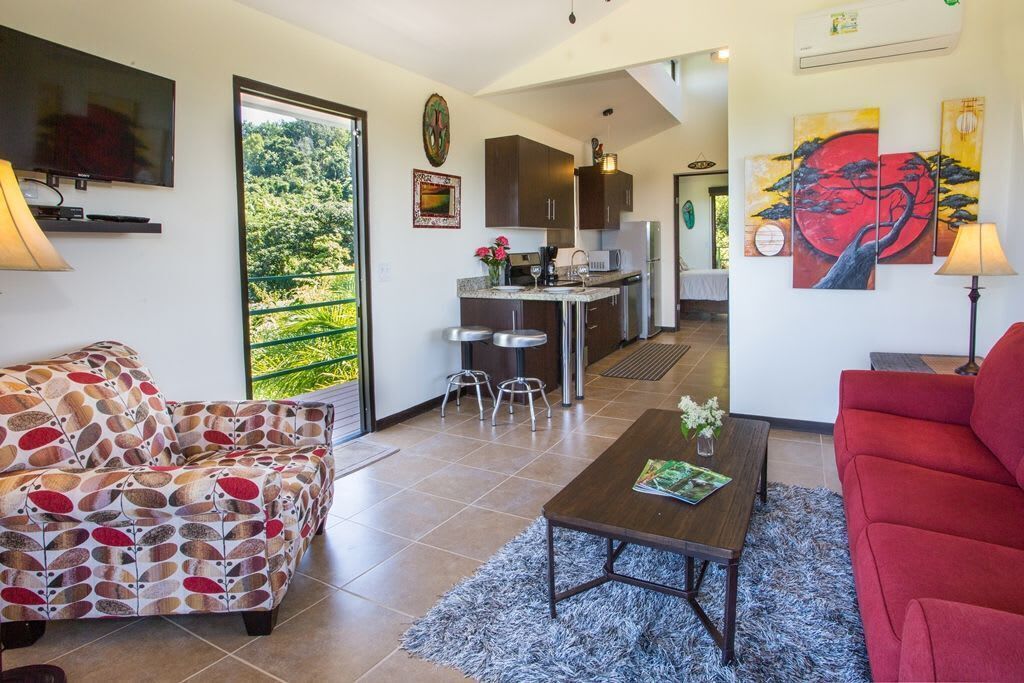 A modern living room with a sofa, a coffee table, and a red couch. There is a kitchen area with cabinets and a dining table.