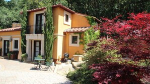 Una villa amarilla con techo de tejas, balcón y patio empedrado con mobiliario exterior.
