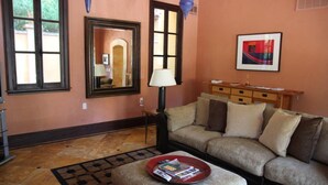Una sala de estar con un sofá, una mesa de centro con revistas, una lámpara, un cuadro enmarcado y una ventana con vistas.