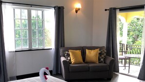 Ein Hotelzimmer mit einem großen Bett, einem grauen Sofa mit gelben Kissen, einer Wandlampe und einem Balkon mit Tisch und Stühlen.
