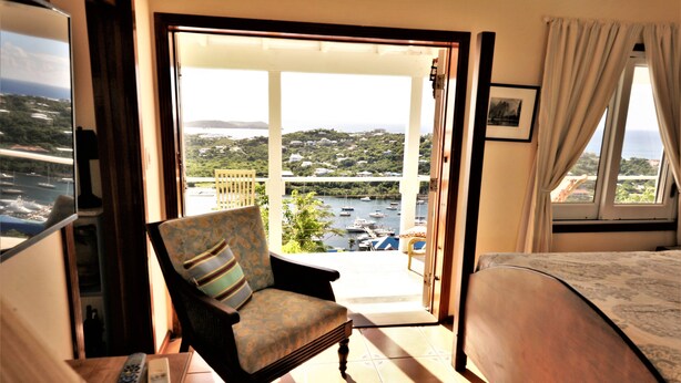 A room with a bed, a chair, a balcony with a view of boats and greenery, and a remote control on the floor. - The 'Cottage' at Lion's Gate - Check for our Spring & Summer Specials (Red Hook)