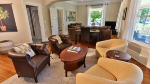 Una sala de estar con un sofá de cuero marrón, un sillón beige y una mesa de centro de madera. Se ve una zona de comedor con una mesa y sillas al fondo.