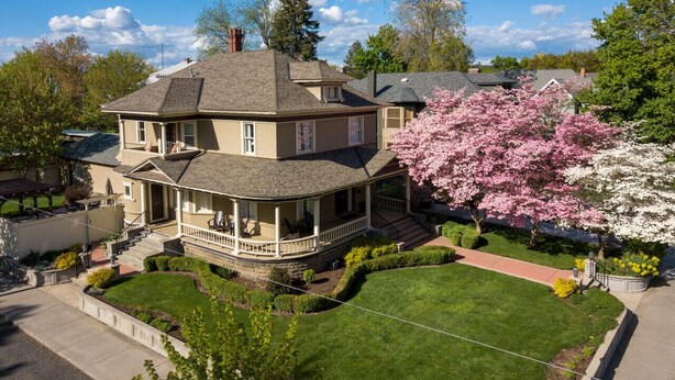 Una casa de dos pisos con un porche que rodea toda la estructura, un césped bien cuidado y un árbol grande con flores.