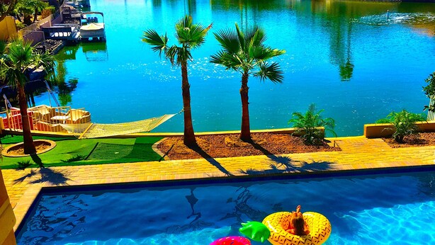 A swimming pool with two people in inflatable rings, palm trees, and a boat docked in the background. - NEW! Luxury Lakefront Estate with Pool and Putting Greens (Glendale)