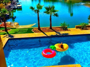 A swimming pool with two people in inflatable rings, palm trees, and a boat docked in the background.