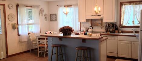 A spacious kitchen with a central island, wooden bar stools, a chandelier, and a view of the beach through the window.