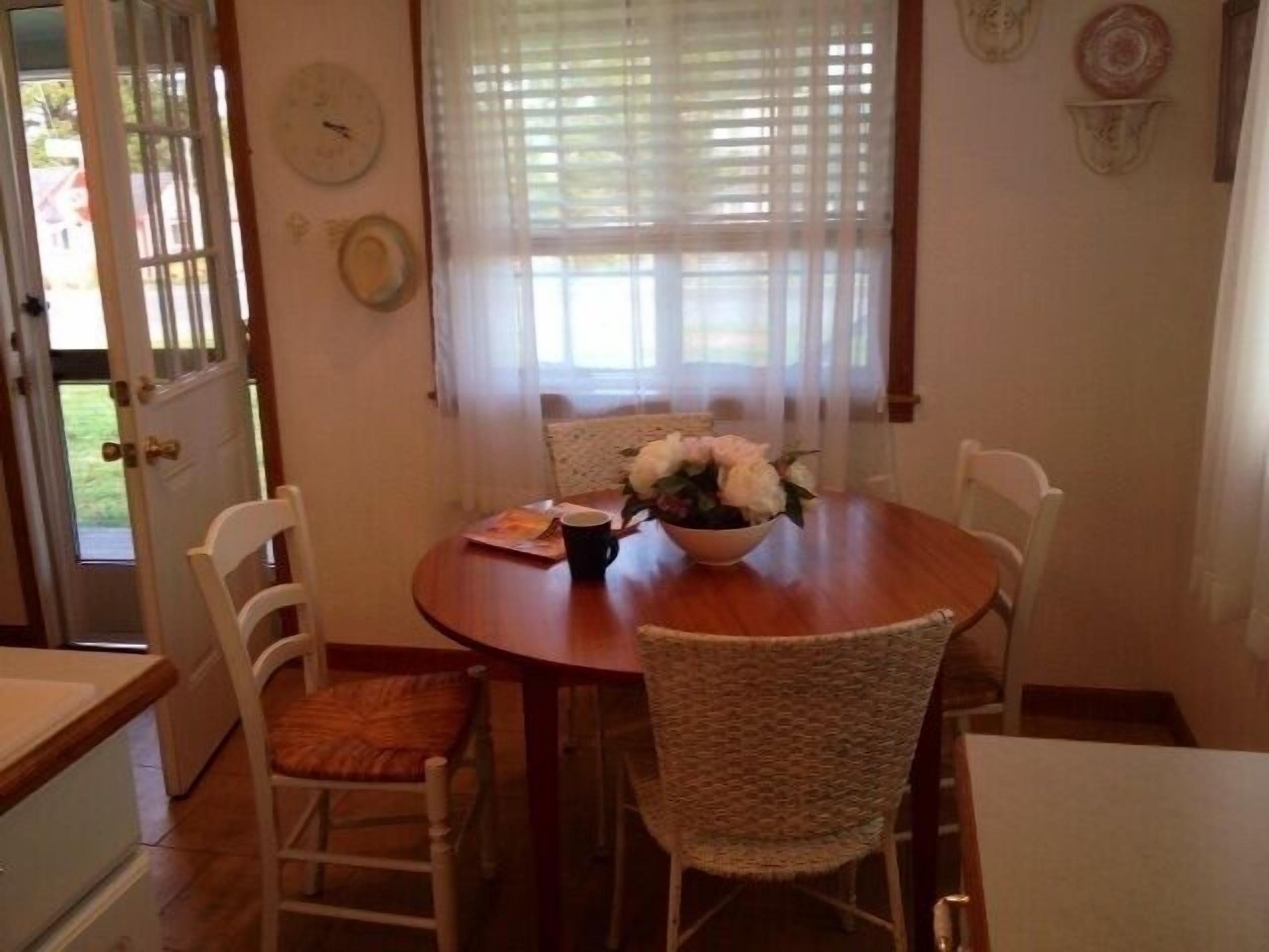 Un comedor con una mesa redonda de madera, sillas blancas, un cuenco de flores y una puerta con ventanal.