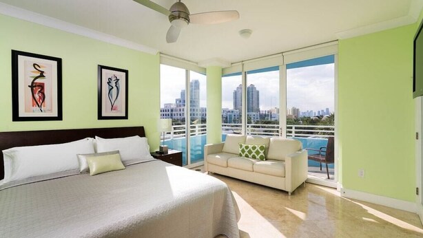 A hotel room with a bed, a sofa, a ceiling fan, and large windows offering a city view. - Incredible Ocean Views! Luxury 2BD in Heart of South Beach & Steps to the Sand! (Miami Beach)