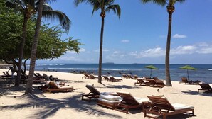 A beach with rows of wooden lounge chairs and palm trees, a clear blue sky, and the ocean in the background.