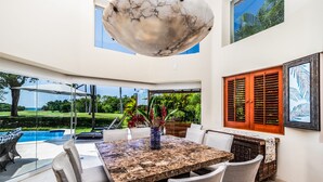 A spacious dining area with a marble table and comfortable chairs, overlooking a pool and lush greenery.