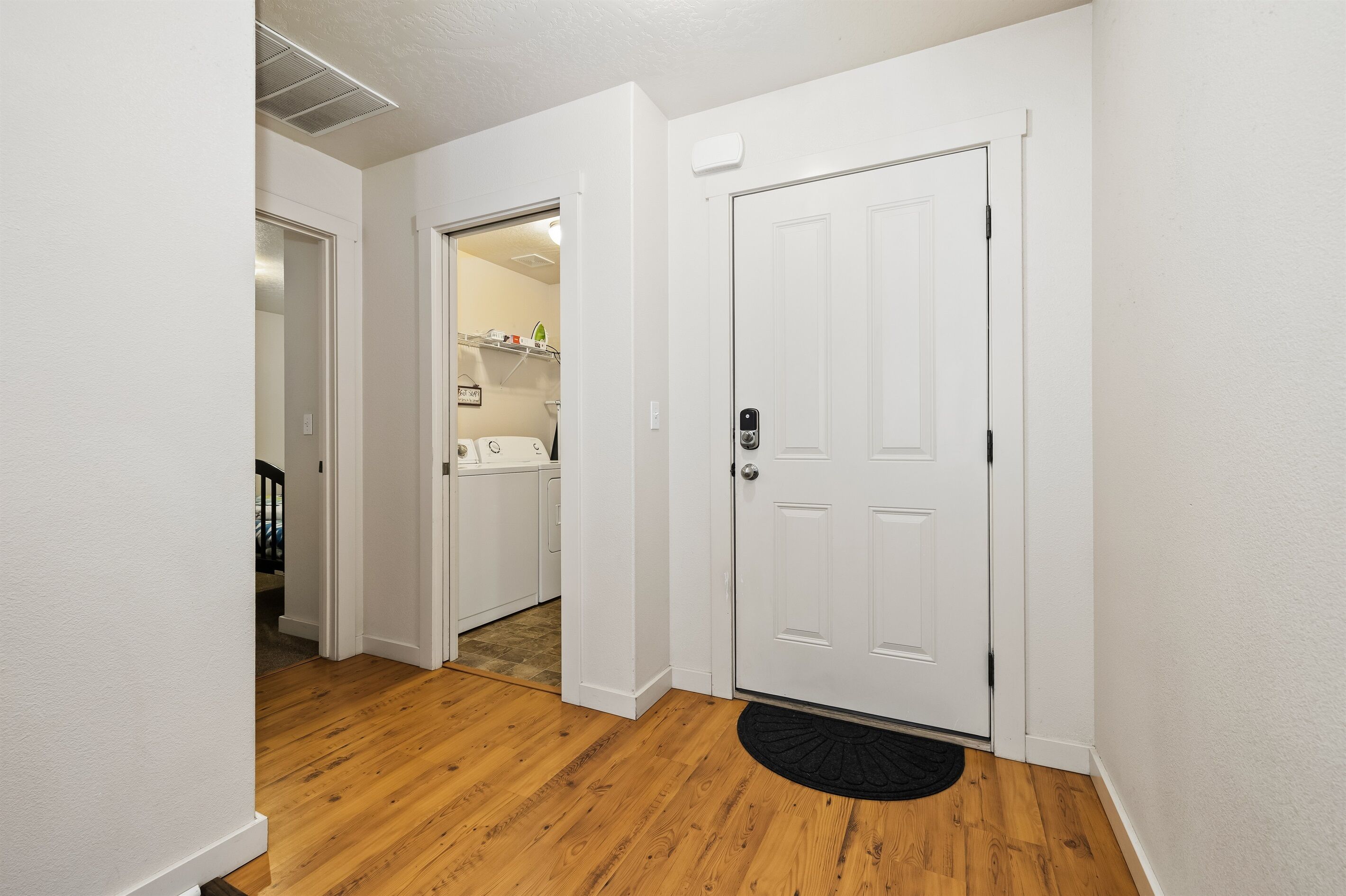 Un couloir doté d’un plancher en bois, une porte blanche avec un paillasson noir, et une salle de bain visible, équipée d’une douche et de toilettes.