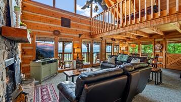 A spacious log cabin interior with a fireplace, leather sofa, dining area, and large windows offering scenic views.