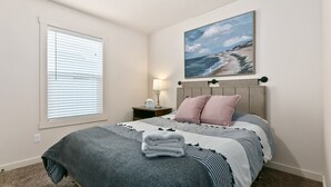 A bedroom with a bed, a nightstand, a lamp, and a painting of a beach scene. - Fenced Yard | Open Concept | Near The Village (Meridian)