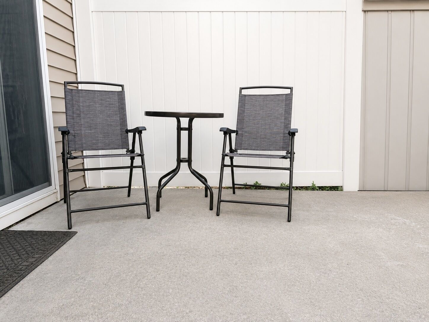 Une terrasse avec deux chaises et une table, une porte-fenêtre coulissante et une clôture blanche.