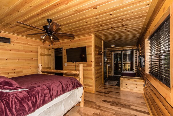 A wooden cabin room with a bed, a ceiling fan, a television, and a window with blinds.