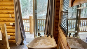 Un baño de cabaña de madera con una bañera grande llena de burbujas, un banco de madera con toallas y una ventana con cortinas.