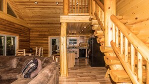 Interior de una cabaña de troncos con una escalera de madera, una sala de estar con sofá y mesa de centro, y una zona de cocina.
