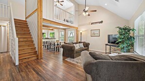 A spacious living area with a wooden staircase, a ceiling fan, a large plant, and a television. - Riverfront Retreat w/ Decks & Views (Baytown)