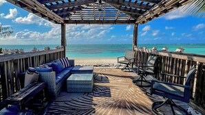A wooden deck with a sofa, chairs, and a patterned rug overlooking a beach. - Private Beachfront Luxury Estate pool with waterfall free concierge – Sleeps 16 (North Caicos)
