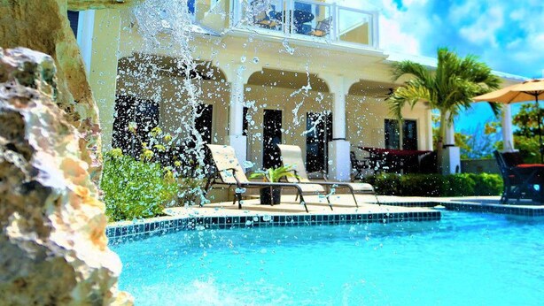 A poolside view of a resort with a clear blue pool, lounge chairs, and a building with a balcony overlooking the pool. - Private Beachfront Luxury Estate pool with waterfall free concierge – Sleeps 16 (North Caicos)