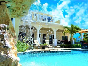 A poolside view of a resort with a clear blue pool, lounge chairs, and a building with a balcony overlooking the pool.