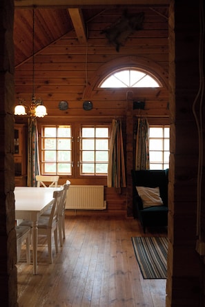 A wooden interior with a dining table, chairs, a sofa, and a window with curtains. - Log cabin for 4 + 2 people, 80m2 in the middle of the forest between 2 lakes (Hallaryd)