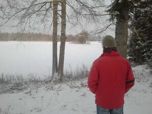 A person standing in a snowy outdoor area with trees and a body of water in the background.